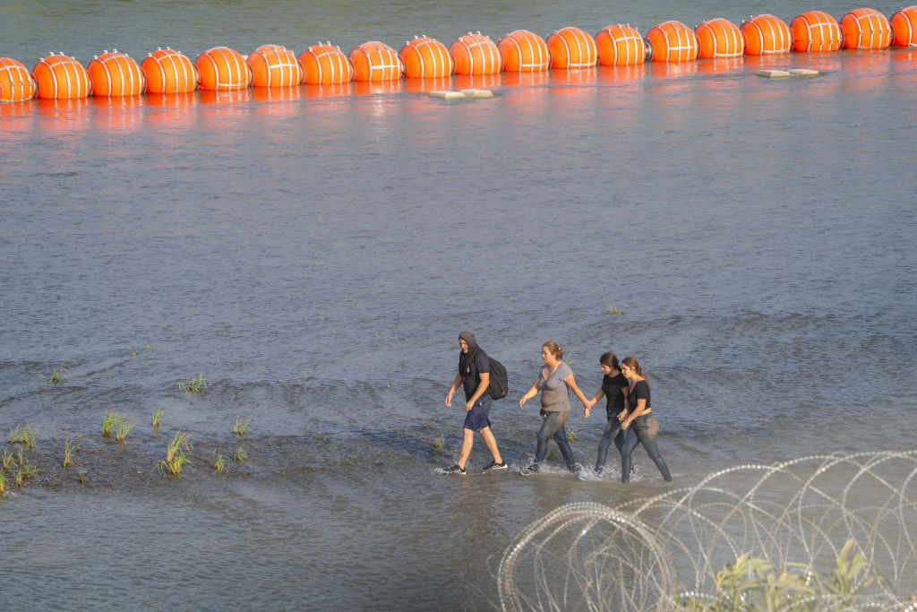 Migrantes caminan entre alambre de concertina y una cadena de boyas colocadas en el agua a lo largo de la frontera del Rio Grande con Mexico en Eagle Pass, Texas.